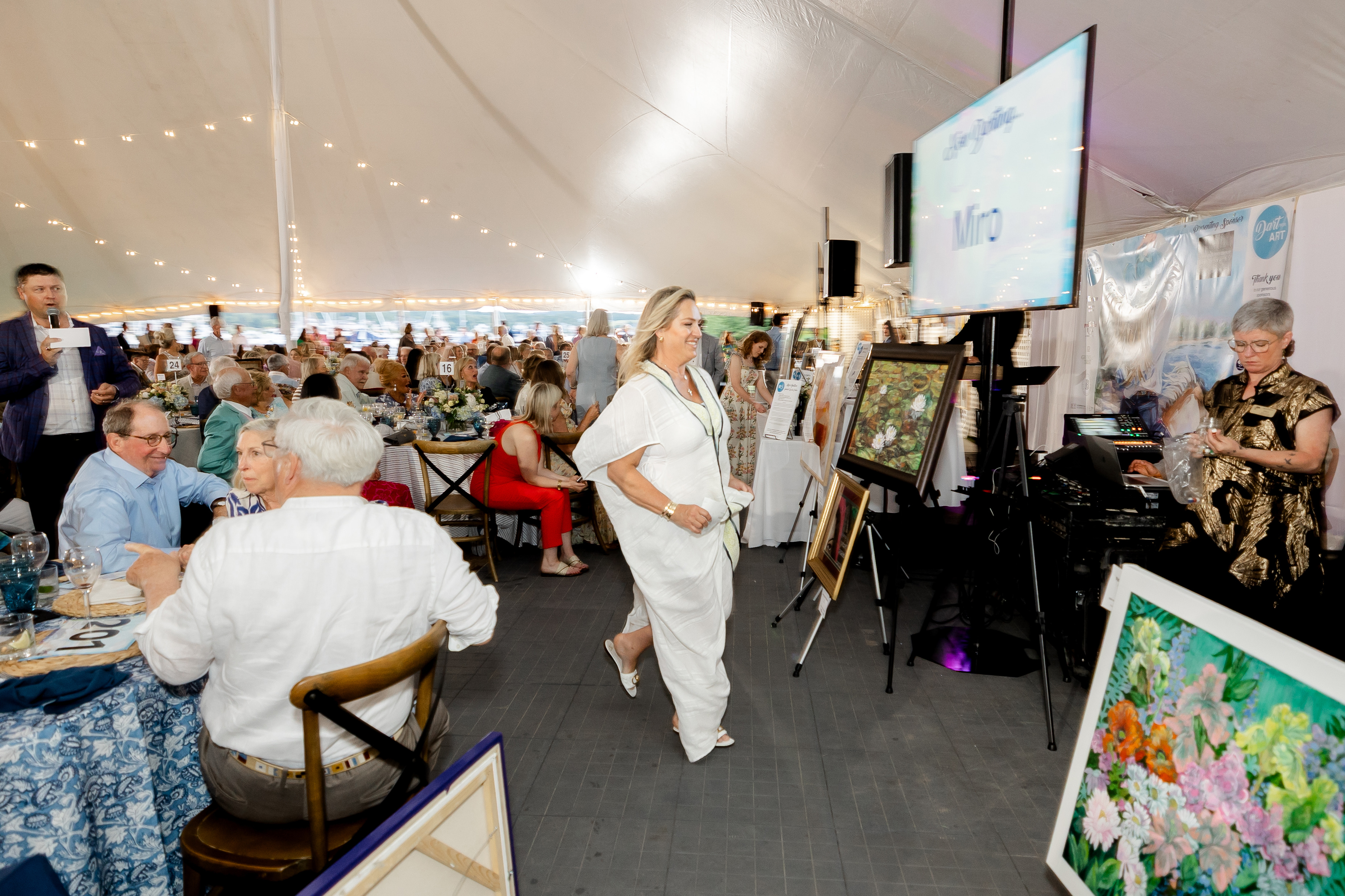 A blonde woman in a white outfit smiling as she races toward art on a gallery wall during an event