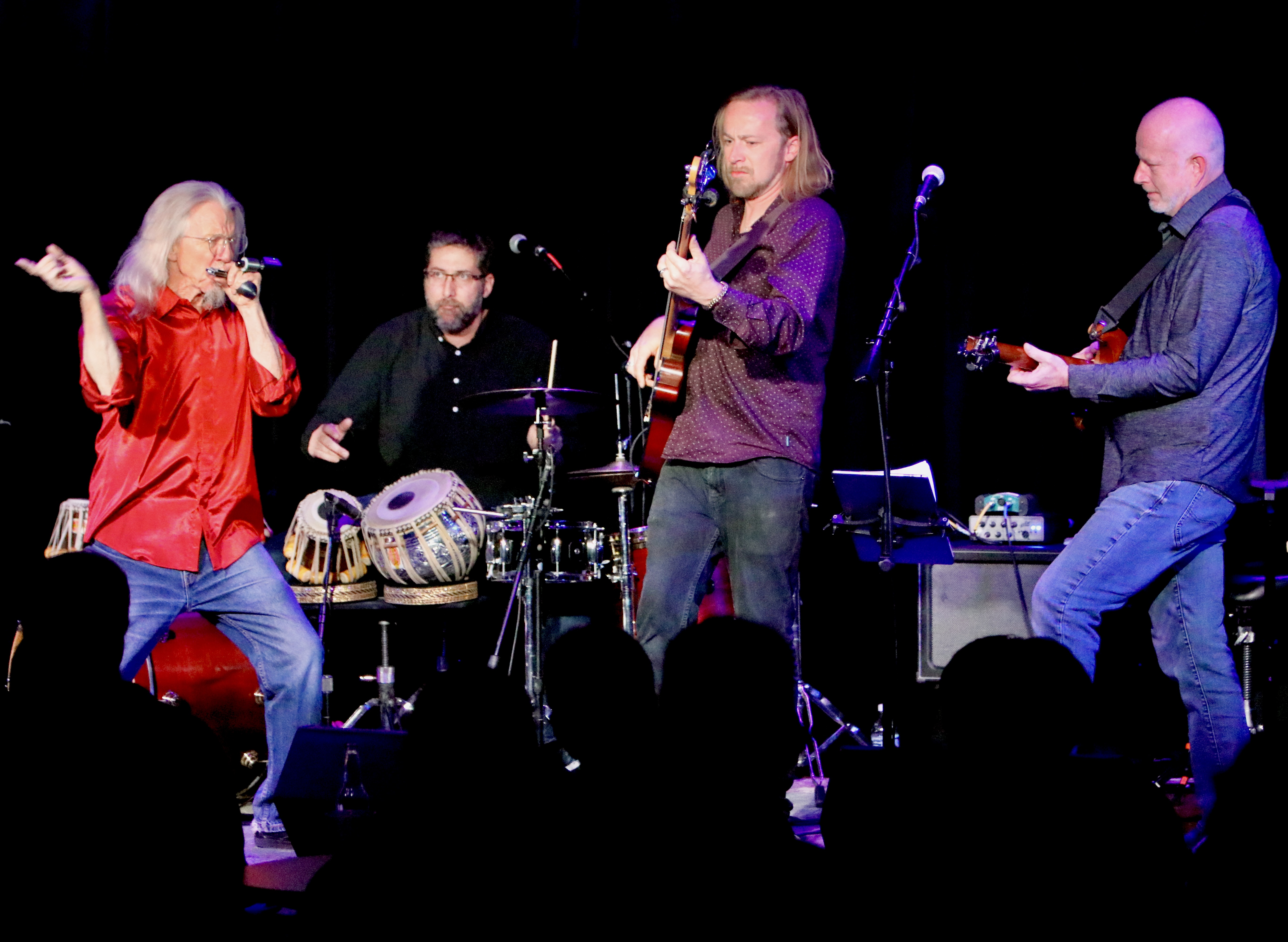 A band performs on stage with four musicians. One sings into a microphone wearing a bright red shirt, another plays the tabla drums, and two others play guitars. Stage lights highlight them against a dark background while the audience watches in silhouette.