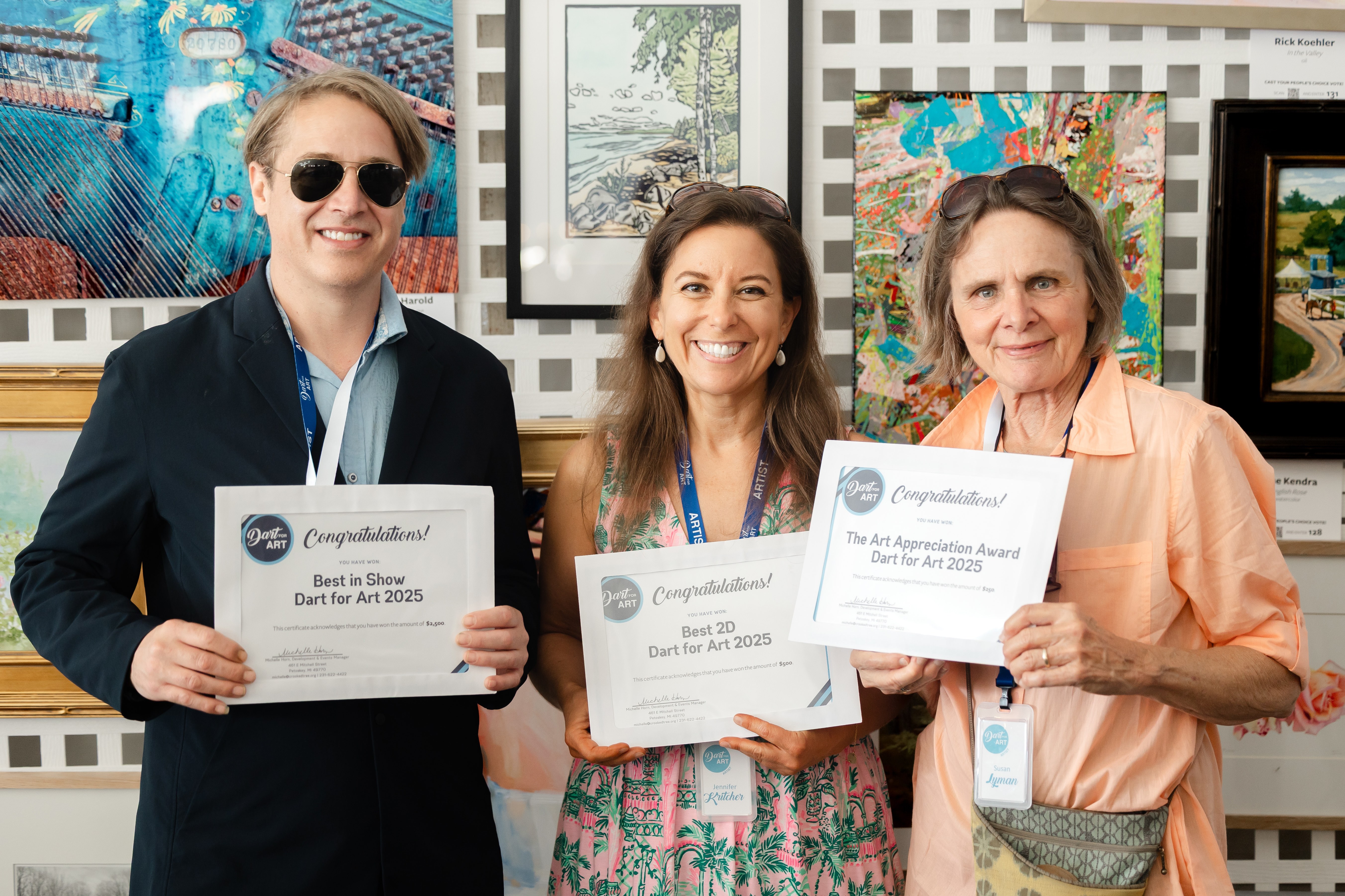 A man and two women stand smiling with their award certificates in front of the art gallery wall