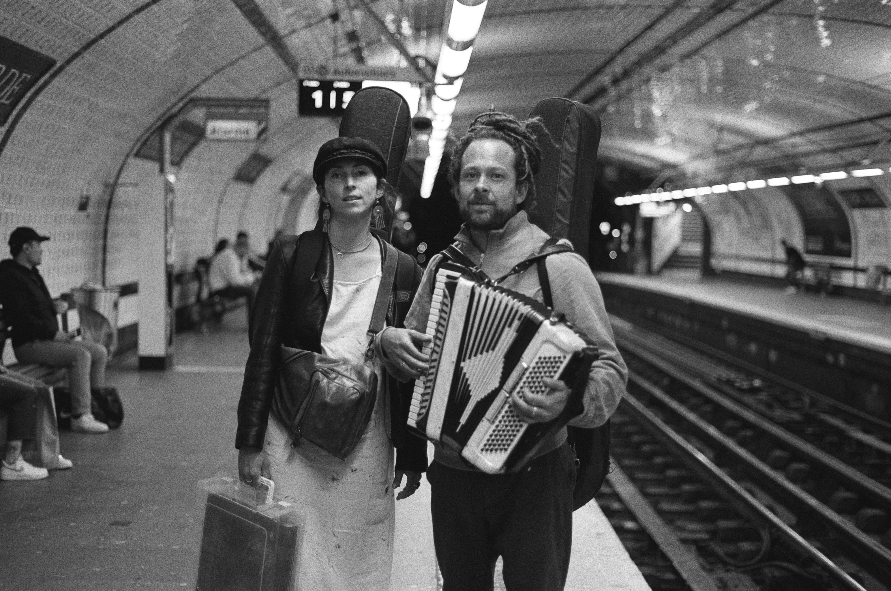 : A man and woman stand on a subway platform with musical instruments. The woman carries a case and wears a leather jacket and cap, while the man holds an accordion with a guitar strapped to his back. The train tracks and tiled station walls stretch into the distance.