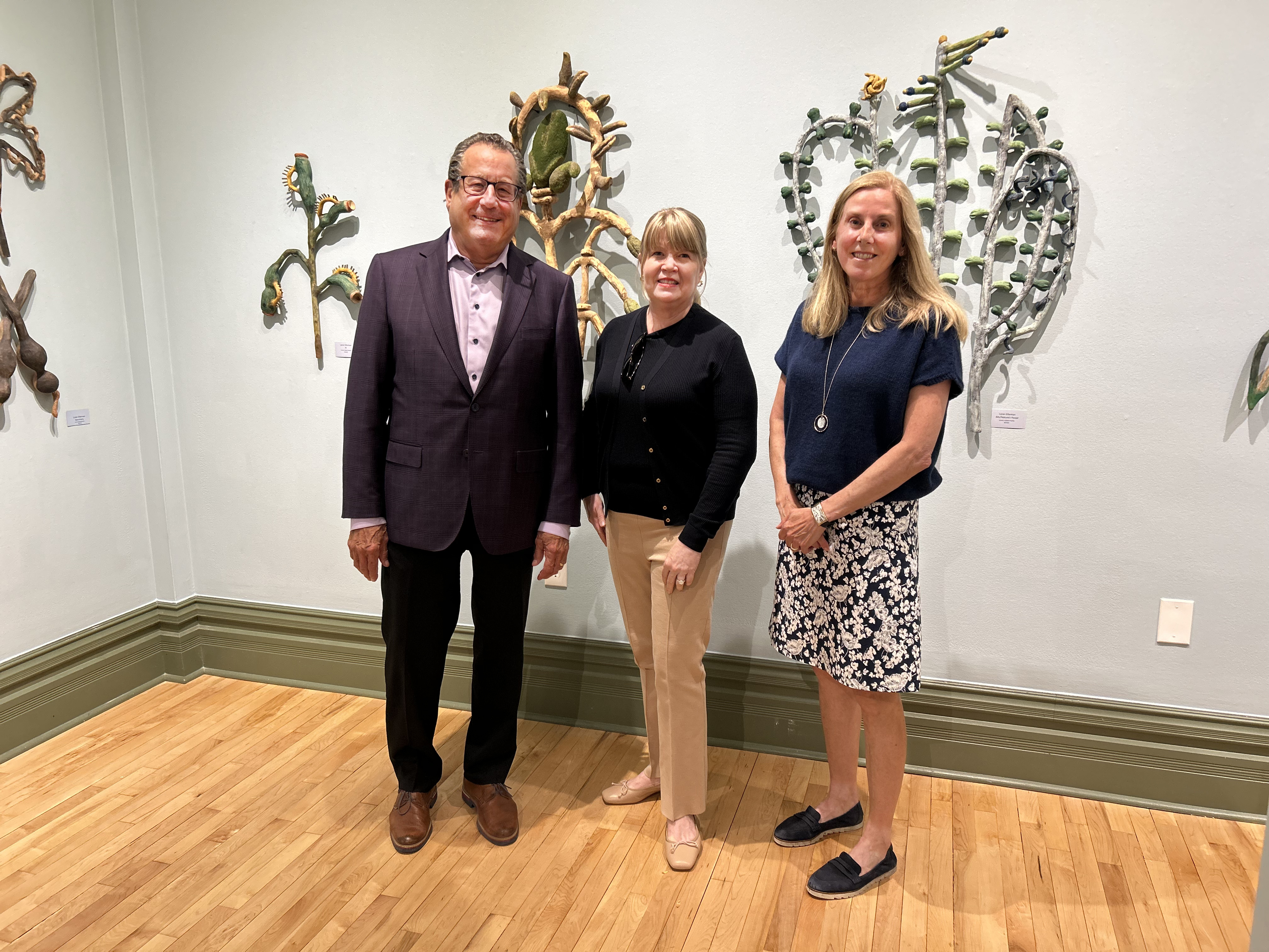 Three people stand together in front of a wall displaying sculptural artworks shaped like plants and leaves. The man on the left wears a suit jacket, while the two women beside him wear casual tops with slacks and a skirt, all smiling for the photo.