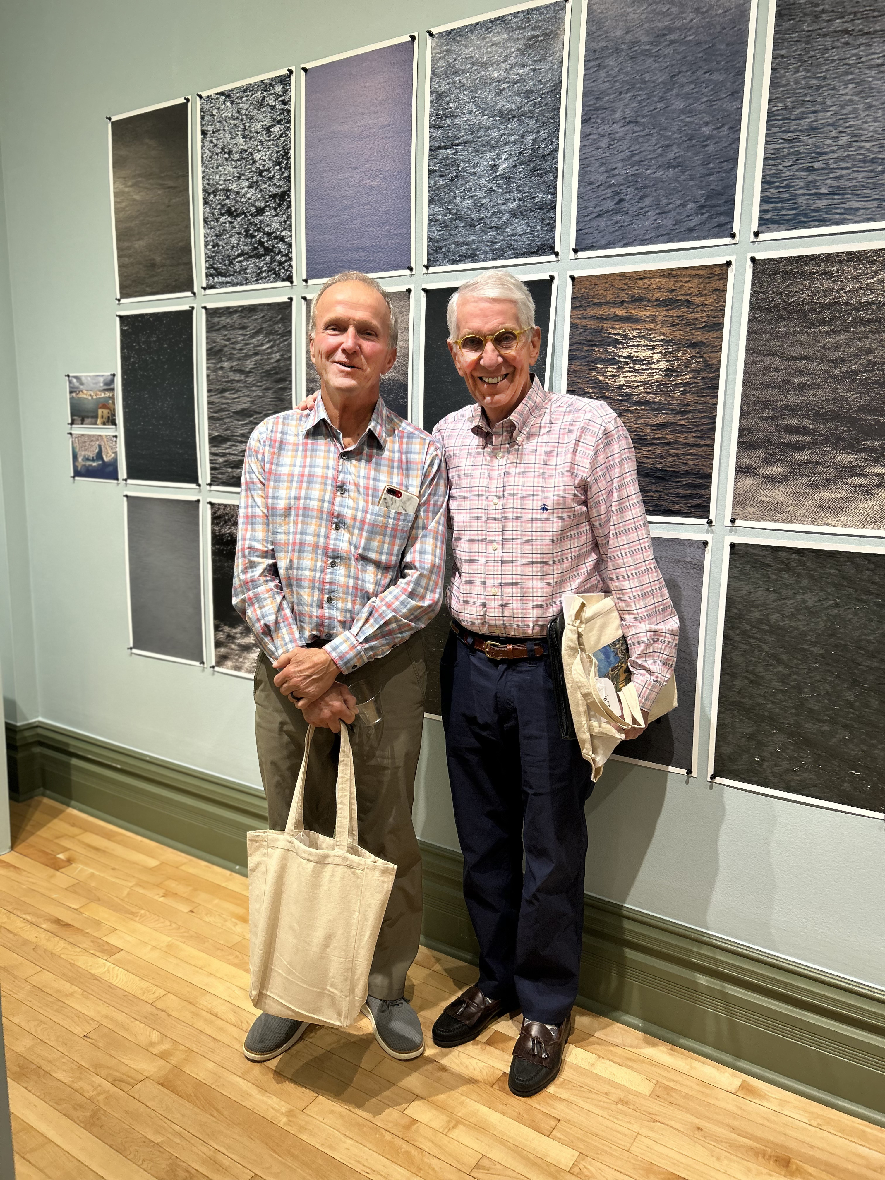 Two men stand side by side in an art gallery, smiling in front of a grid of photographs depicting different views of water. Both are wearing plaid shirts, and one holds a canvas tote bag while the other carries folded materials in his hand.