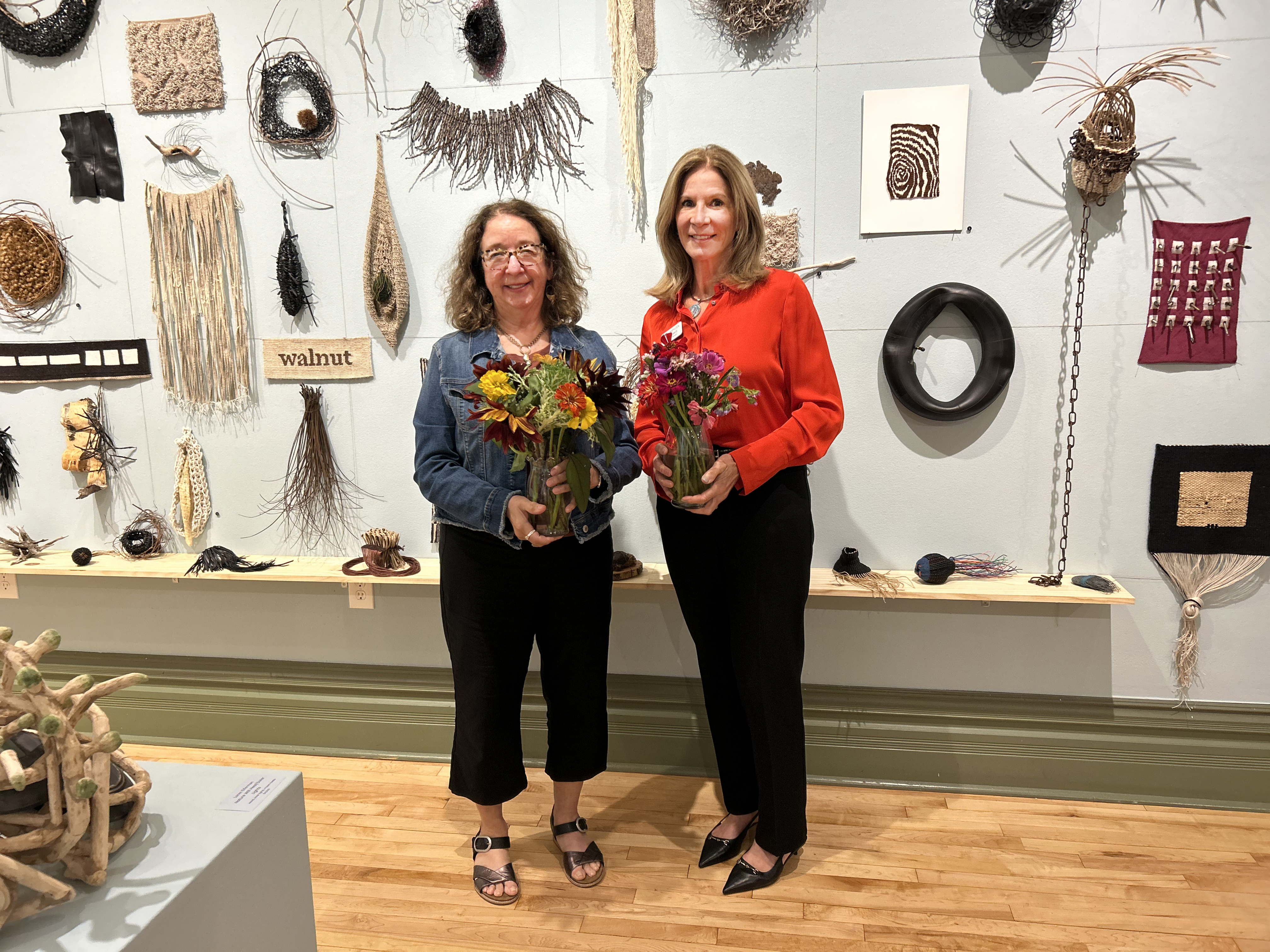 Two women pose in front of a wall display featuring mixed-media fiber artworks and natural materials. Each is holding a bouquet of flowers—one wearing a denim jacket and sandals, the other in a bright red blouse and black pants.