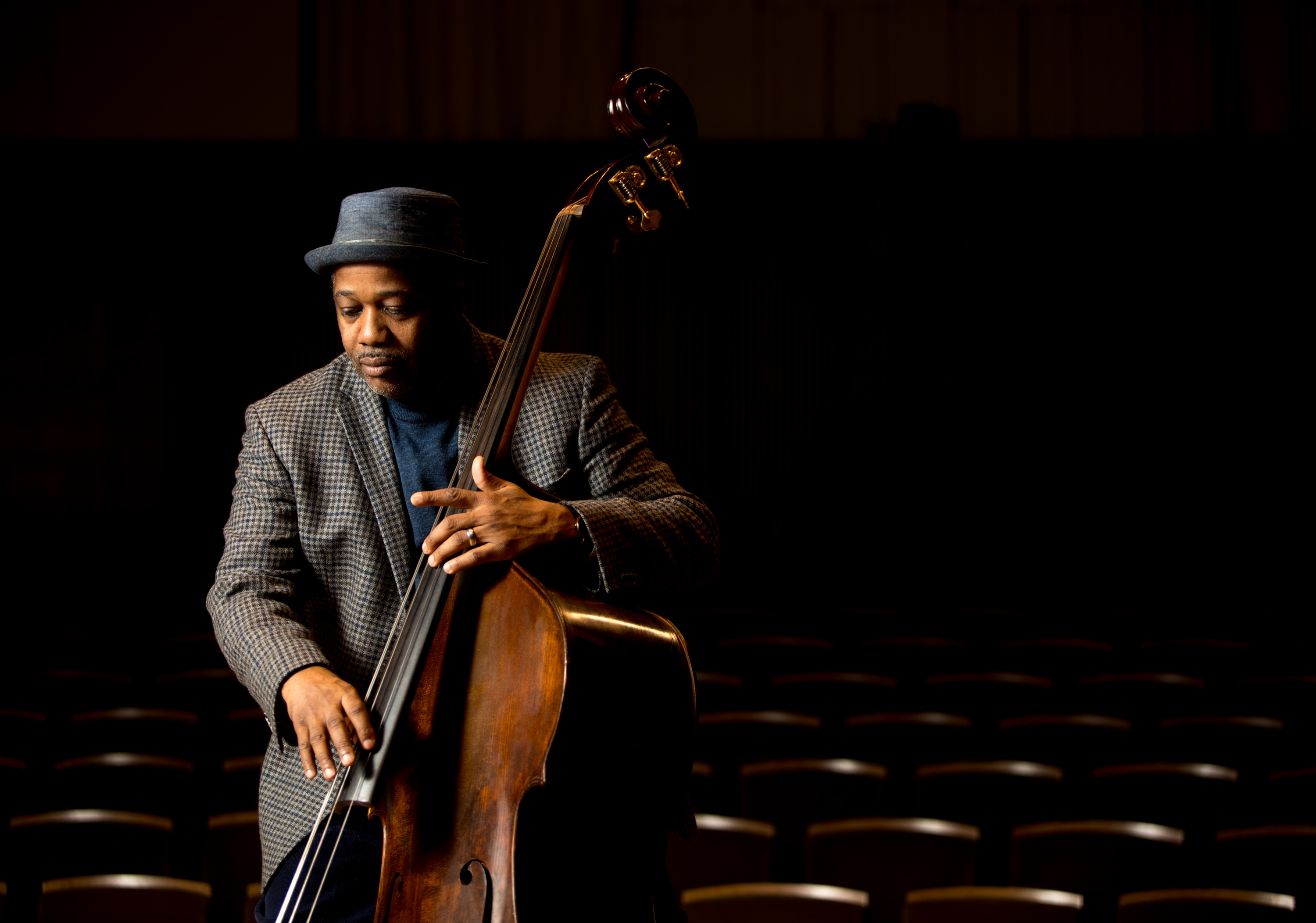 A musician in a hat and checkered jacket plays an upright bass on stage. The empty rows of seats behind him fade into darkness as a spotlight highlights his focused expression and hands on the instrument.